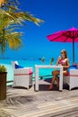 Beautiful woman in bikini is sitting in beach bar on the Maldives sandy beach and holding colorful fruity drink. Luxury travel.