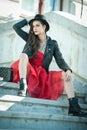 Beautiful woman with black hat, red dress and boots posing sitting on stairs. Young brunette spending time during autumn Royalty Free Stock Photo