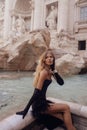 beautiful woman with blond curly hair wearing elegant black dress and accessories, posing in front of a fountain di Trevi, famous Royalty Free Stock Photo