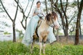 Beautiful woman with blue dress stay on white horse and stand in field near village during early morning also look at camera