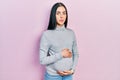 Beautiful woman with blue eyes expecting a baby, touching pregnant belly relaxed with serious expression on face Royalty Free Stock Photo