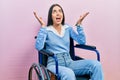 Beautiful woman with blue eyes sitting on wheelchair celebrating mad and crazy for success with arms raised and closed eyes