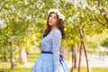 A beautiful woman in a blue puffy dress with a rim of white flowers on her head walks through the park between blooming apple tree