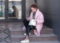 Beautiful woman with bundled hair, purse in pink coat and black pants sitting on stairs and using cell phone