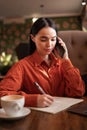 Beautiful woman in cafe, drinking coffee, writing in notebook, checking documents on lunch break, call for client on Royalty Free Stock Photo