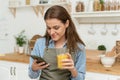 Beautiful woman cooking vegetables in cozy wooden kitchen while looking recipe on smartphone and drinking juice. Young