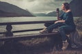 Beautiful woman with cup of coffee or tea sitting on a banch near the wild lake, with mountains on the background.