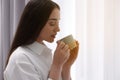 Beautiful woman with cup of drink near window at home, space for text. Lazy morning Royalty Free Stock Photo