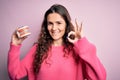 Beautiful woman with curly hair holding plastic teeth with dental braces over pink background doing ok sign with fingers, Royalty Free Stock Photo