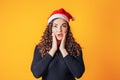 Beautiful woman with curly hair and santa hat on her head. The woman shows emotion of surprise and touched her face with her hands