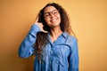 Beautiful woman with curly hair wearing casual denim shirt and glasses over yellow background smiling doing phone gesture with Royalty Free Stock Photo