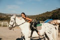 Beautiful woman with dark hair in elegant sportive suit posing near white horse in stable Royalty Free Stock Photo