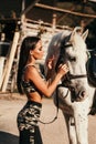 Beautiful woman with dark hair in elegant sportive suit posing near white horse in stable Royalty Free Stock Photo