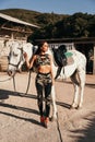 Beautiful woman with dark hair in elegant sportive suit posing near white horse in stable Royalty Free Stock Photo