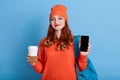 Beautiful woman dressed orange jumper and cap, carries takeaway coffee, isolated over blue wall, holding smart phone with blank Royalty Free Stock Photo