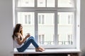 Beautiful young woman drinking coffee and looking through window while sitting at windowsill at home Royalty Free Stock Photo