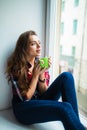 Beautiful young woman drinking coffee and looking through window while sitting at windowsill at home Royalty Free Stock Photo