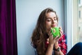 Beautiful young woman drinking coffee and looking through window while sitting at windowsill at home Royalty Free Stock Photo