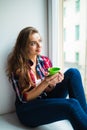 Beautiful young woman drinking coffee and looking through window while sitting at windowsill at home Royalty Free Stock Photo