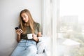 Beautiful young woman drinking coffee use mobile phone and looking through window while sitting at windowsill at home Royalty Free Stock Photo