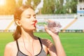 Beautiful woman is drinking water and listening to the music on headphones at the stadium. Girl is having a break after training Royalty Free Stock Photo