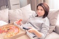 Surprised woman eating pizza and watching TV with remote control at home, warm tone Royalty Free Stock Photo