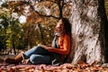 A beautiful woman enjoying the sun in a park in an autumnal day. She is sitting and leaning against a tree while having a book on