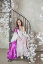 Beautiful woman in fantasy white and purple rococo style medieval dress sitting on the stairway with pink flowers