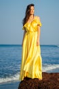 Beautiful woman with flying long butterfly silk yellow dress posing on beach standing on a rock