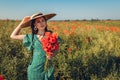 Beautiful woman gathered bouquet of poppies flowers walking in summer field. Stylish girl wearing vintage straw hat