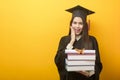 Beautiful woman in graduation gown is holding books and certificate on yellow background Royalty Free Stock Photo