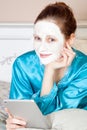 Beautiful woman with green nightie and white facial mask resting on bed and holding tablet before sleeping.