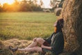 Beautiful woman in hat enjoying sunset, sitting at haystacks in evening summer field. Tranquility