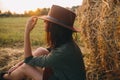 Beautiful woman in hat enjoying sunset, sitting at haystacks in evening summer field. Tranquility