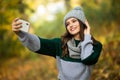 Beautiful young woman in hat, scarf and jacket in autumn takes a selfie on her phone in autumn park Royalty Free Stock Photo