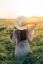 Beautiful woman in hat with wildflowers bouquet standing in barley field in sunset light. Stylish female relaxing in evening