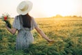 Beautiful woman in hat with wildflowers bouquet standing in barley field in sunset light. Stylish female relaxing in evening