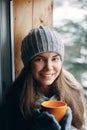 Beautiful  woman holding and drinking a cup of coffee or cocoa in gloves sitting home by the window. Blurred winter snow tree back Royalty Free Stock Photo