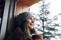 Beautiful  woman holding and drinking a cup of coffee or cocoa in gloves sitting home by the window. Blurred winter snow tree back Royalty Free Stock Photo