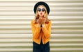 Beautiful woman holding hands blowing red lips sending sweet air kiss wearing yellow jacket, black round hat on wall Royalty Free Stock Photo