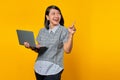 Beautiful woman holding laptop looking sideways and pointing with finger at empty space on yellow background