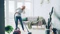 Beautiful woman is hoovering the floor at home using modern vacuum cleaner and listening to music with headphones Royalty Free Stock Photo