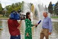 A beautiful woman journalist in a green dress with a microphone and a cameraman with a video camera is interviewing a middle-aged