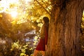 Beautiful woman leaning on a tree quietly watching the sun and the autumn landscape. Young woman enjoying nature in autumn. Autumn