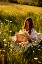 a beautiful woman in a light dress sits in a field of daisies at sunset holding a hat in her hands Royalty Free Stock Photo