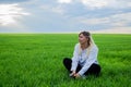 A beautiful woman listens to music in headphones while sitting on a green field against a cloudy sky. Pretty woman looks