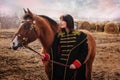 A beautiful woman with long and black hair in a historical hussar costume stands in a field with a horse Royalty Free Stock Photo