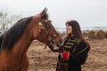 A beautiful woman with long and black hair in a historical hussar costume stands in a field with a horse Royalty Free Stock Photo