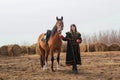 A beautiful woman with long and black hair in a historical hussar costume stands in a field with a horse Royalty Free Stock Photo