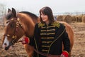 A beautiful woman with long and black hair in a historical hussar costume stands in a field with a horse Royalty Free Stock Photo